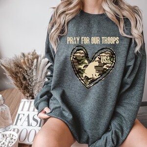 K&ouml;nnte beinhalten: Ein graues Sweatshirt mit dem Text "Pray for our Troops" und einem Tarnmusterherz mit dem Text "USA" darin.
