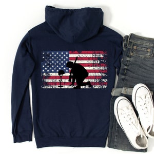 Puede incluir: Sudadera con capucha azul marino con un dise&ntilde;o de bandera estadounidense desgastado que presenta la silueta de un soldado arrodillado sosteniendo un rifle. El soldado est&aacute; arrodillado frente a un casco y un ramo de flores.