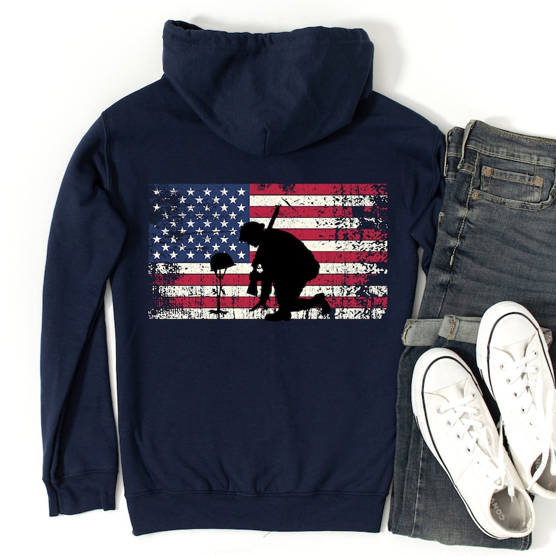 Puede incluir: Sudadera con capucha azul marino con un dise&ntilde;o de bandera estadounidense desgastado que presenta la silueta de un soldado arrodillado sosteniendo un rifle. El soldado est&aacute; arrodillado frente a un casco y un ramo de flores.
