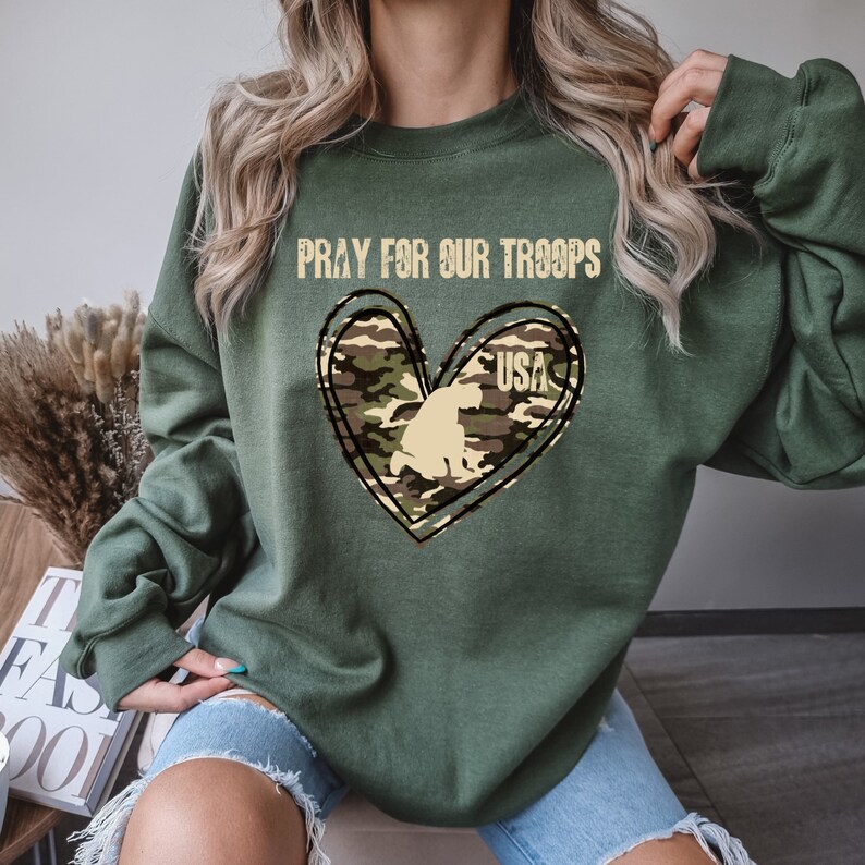 K&ouml;nnte beinhalten: Gr&uuml;nes Sweatshirt mit einem Tarnmuster-Herzdesign und dem aufgedruckten Text "Pray for our Troops" und "USA".
