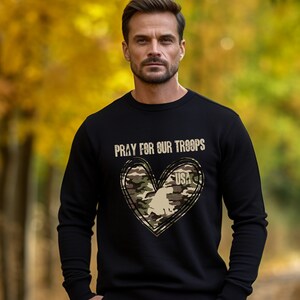 K&ouml;nnte beinhalten: Schwarzes Sweatshirt mit einem Tarnmuster-Herzdesign und dem Text "Pray for our Troops" und "USA".