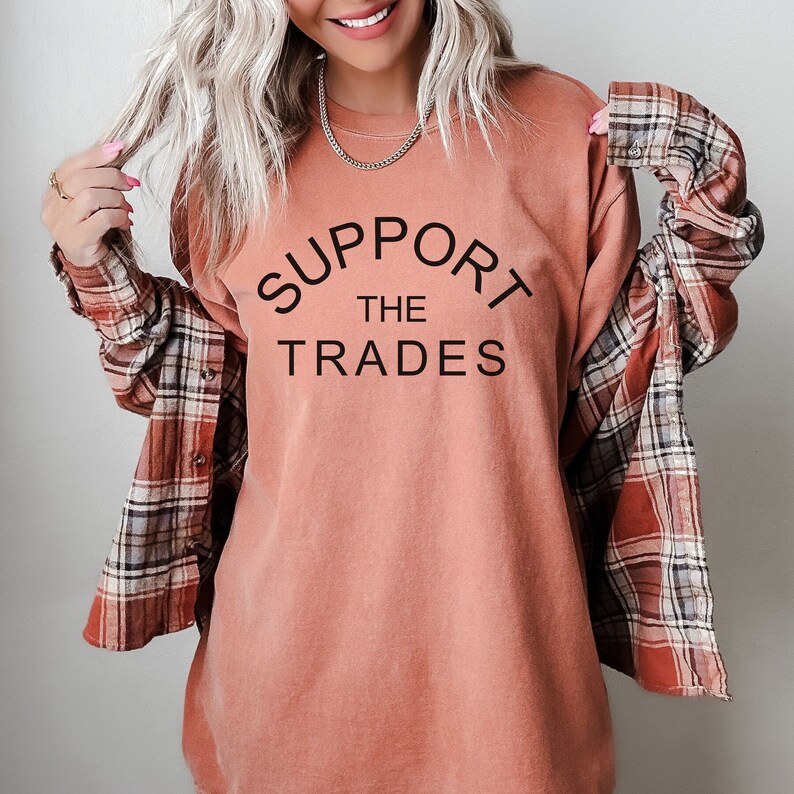 May include: A woman wearing a peach colored long-sleeved shirt with the text "SUPPORT THE TRADES" printed on the front. She is wearing a plaid shirt over the top.