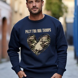 K&ouml;nnte beinhalten: Marineblauer Pullover mit einem Tarnmuster-Herzdesign und dem Text "Pray for our Troops" und "USA".
