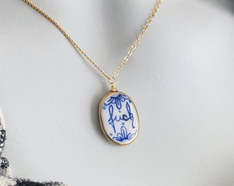 Pode incluir: Um colar de corrente dourada com um pingente oval de superfície cerâmica branca, decorado com padrões florais azuis e a palavra "fuck" escrita em letra cursiva. O pingente é emoldurado por uma fina armação de metal de cor dourada.
