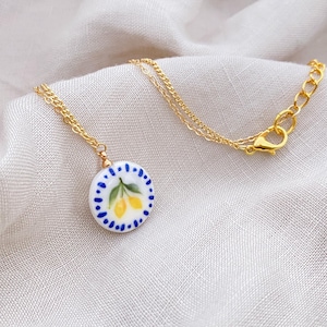 Op de afbeelding: Een goudkleurige ketting met een ronde, witte keramische hanger. De hanger is versierd met een handgeschilderd ontwerp van twee gele citroenen met groene bladeren, omringd door een rand van kleine, blauwe geschilderde stippen.