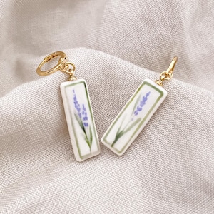 Può includere: Orecchini color oro con pendenti rettangolari bianchi. Ogni pendente presenta un design a rametto di lavanda con fiori viola e foglie verdi. Gli orecchini sono su uno sfondo di lino neutro.