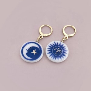 Puede incluir: Un par de pendientes de temática celestial. Cada pendiente presenta un colgante redondo de cerámica blanca con un diseño azul. Uno representa una luna creciente y una estrella, el otro un sol con una cara. Cierres dorados.