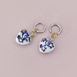 May include: A pair of heart-shaped earrings. Each earring features a white ceramic heart with a blue crescent moon and gold star design. The earrings have silver hoops and gold-colored connectors.