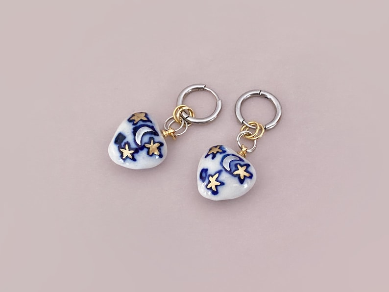 May include: A pair of heart-shaped earrings. Each earring features a white ceramic heart with a blue crescent moon and gold star design. The earrings have silver hoops and gold-colored connectors.
