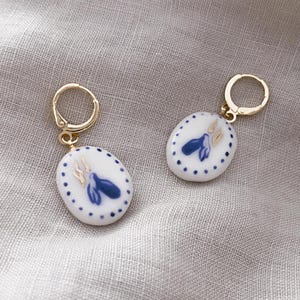 Könnte beinhalten: Ein Paar goldfarbene Klappcreolen. Jeder Ohrring hat einen weißen, ovalen Anhänger mit blauem Blumendesign und goldenen Akzenten. Der Anhänger ist von kleinen blauen Punkten umgeben. Auf neutralem Leinenhintergrund.
