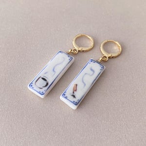 Op de afbeelding: Een paar rechthoekige oorbellen met goudkleurige creolen. De witte keramische rechthoeken hebben een blauwe rand en een minimalistisch ontwerp met een kopje en een vogel. De oorbellen staan tegen een neutrale achtergrond.