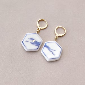Puede incluir: Un par de pendientes hexagonales con un diseño blanco y azul. Los pendientes tienen aros dorados y un pequeño acento dorado en la superficie blanca. Un accesorio único.