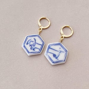 May include: Gold-coloured hoop earrings with hexagonal white ceramic pendants. Each pendant has a blue dachshund illustration within a blue border. These earrings are a unique, artistic accessory.