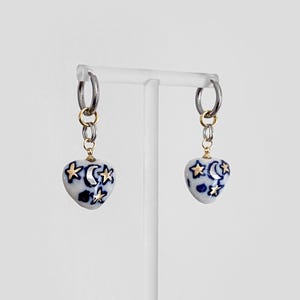 May include: A pair of heart-shaped earrings with a celestial design. The white ceramic hearts feature blue patterns and gold stars and moons. The earrings hang from silver hoops with gold accents.