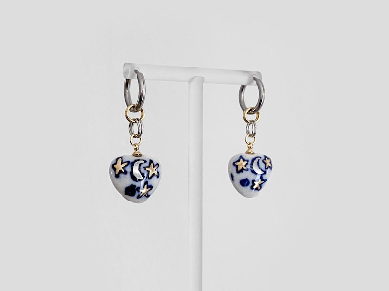 May include: A pair of heart-shaped earrings with a celestial design. The white ceramic hearts feature blue patterns and gold stars and moons. The earrings hang from silver hoops with gold accents.