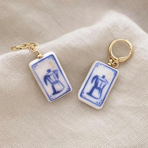 Könnte beinhalten: Ein Paar goldfarbene Creolen mit rechteckigen weißen Keramikanhängern. Jeder Anhänger zeigt eine blau-weiße Illustration einer Kaffeemaschine. Die Ohrringe sind auf einem neutralfarbenen Stoff platziert.