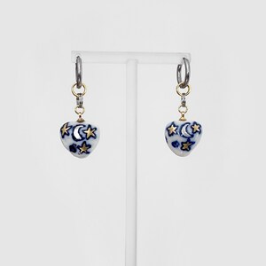 May include: A pair of earrings featuring heart-shaped pendants. The pendants are white with a blue celestial design of moons and stars, accented with gold-colored stars. The earrings hang from silver hoops.