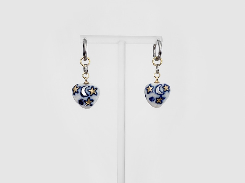 May include: A pair of earrings featuring heart-shaped pendants. The pendants are white with a blue celestial design of moons and stars, accented with gold-colored stars. The earrings hang from silver hoops.