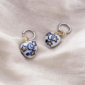 May include: A pair of heart-shaped earrings with a celestial design. The white ceramic earrings feature blue crescent moons and stars, accented with gold details and attached to silver hoops.