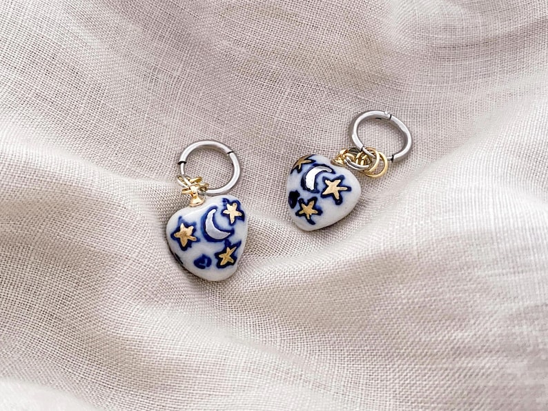 May include: A pair of heart-shaped earrings with a celestial design. The white ceramic earrings feature blue crescent moons and stars, accented with gold details and attached to silver hoops.