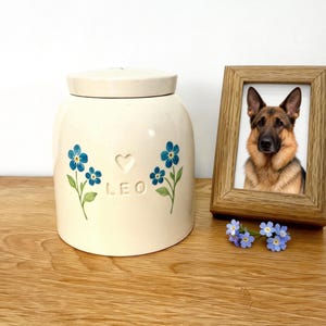 Op de afbeelding: Een crèmekleurige keramische pot met een deksel, versierd met blauwe bloemmotieven en de naam "LEO". Daarnaast een houten fotolijst met een foto van een Duitse herdershond. Een klein boeket blauwe bloemen ligt voor het frame.