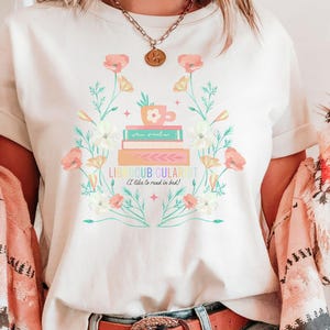 May include: Cream-colored t-shirt featuring a floral design with books, a teacup, and the text "LIBROCUBULARIST (I like to read in bed)". The design includes pink, yellow, and green floral elements. A gold necklace is visible.