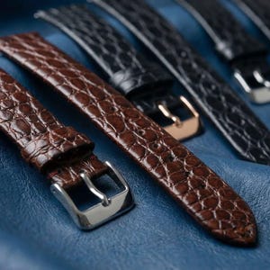 May include: Close-up of several watch straps in various colors. The primary strap is brown with a crocodile pattern and a silver buckle. Other straps are black with silver or gold buckles. The straps are on a blue textured surface.