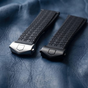 Könnte beinhalten: Zwei schwarze Uhrenarmbänder mit strukturiertem Muster und silbernen und schwarzen Verschlüssen. Die Armbänder werden auf einer blauen Lederoberfläche präsentiert, die die Details des Uhrenzubehörs hervorhebt.