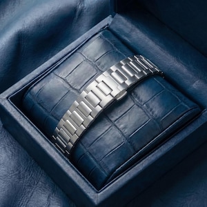 May include: A silver watch strap is displayed on a blue, textured cushion within a matching blue box. The box is open, showing the metallic links and clasp. The background is a darker blue, textured surface.
