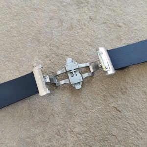 Puede incluir: Una correa de reloj negra con un cierre de mariposa de acero inoxidable. La correa est&aacute; hecha de un material texturizado y flexible. El cierre est&aacute; abierto, mostrando el mecanismo. El fondo es una superficie rugosa de color marr&oacute;n claro.