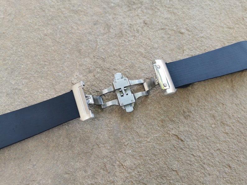 Puede incluir: Una correa de reloj negra con un cierre de mariposa de acero inoxidable. La correa est&aacute; hecha de un material texturizado y flexible. El cierre est&aacute; abierto, mostrando el mecanismo. El fondo es una superficie rugosa de color marr&oacute;n claro.