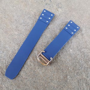 May include: Two navy blue watch straps with silver rivets and a gold-colored buckle. The straps are made of a flexible material and are laid out on a textured, gray surface. The straps are designed to attach to a watch.