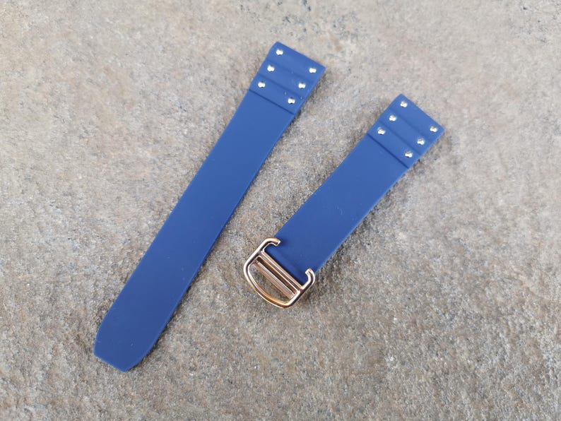 May include: Two navy blue watch straps with silver rivets and a gold-colored buckle. The straps are made of a flexible material and are laid out on a textured, gray surface. The straps are designed to attach to a watch.