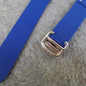 May include: Close-up of a vibrant blue watch strap with a gold-colored buckle. The strap is made of a smooth, flexible material. The buckle has a rectangular design with a polished finish. The strap is designed to be attached to a watch.