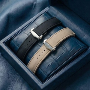 Puede incluir: Una caja de reloj azul con un interior texturizado contiene dos correas de reloj. Una correa es negra, la otra beige. Ambas tienen cierres plateados y se muestran sobre un cojín azul en relieve.