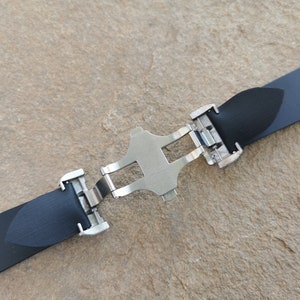 Puede incluir: Una correa de reloj negra con un cierre de mariposa plateado. La correa est&aacute; hecha de un material texturizado y se asegura con un cierre met&aacute;lico. El cierre est&aacute; en el centro de la correa.