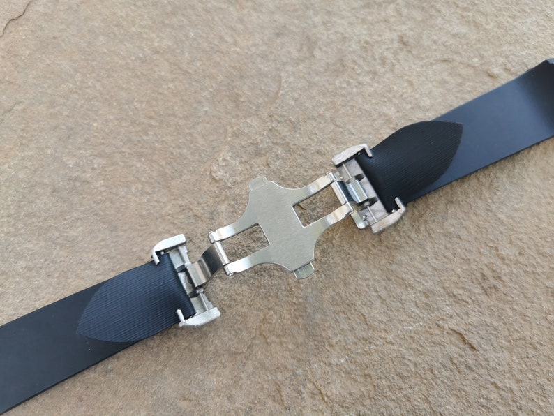 Puede incluir: Una correa de reloj negra con un cierre de mariposa plateado. La correa est&aacute; hecha de un material texturizado y se asegura con un cierre met&aacute;lico. El cierre est&aacute; en el centro de la correa.