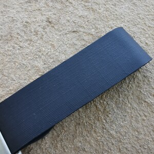 Puede incluir: Primer plano de una correa de cintur&oacute;n de seguridad negra con textura acanalada. La correa est&aacute; en &aacute;ngulo sobre el marco, con una hebilla de metal plateado visible en la parte inferior izquierda. El fondo es una superficie de piedra rugosa de color marr&oacute;n claro.