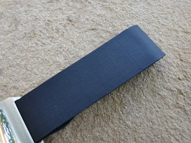 Puede incluir: Primer plano de una correa de cintur&oacute;n de seguridad negra con textura acanalada. La correa est&aacute; en &aacute;ngulo sobre el marco, con una hebilla de metal plateado visible en la parte inferior izquierda. El fondo es una superficie de piedra rugosa de color marr&oacute;n claro.