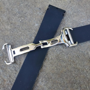 May include: A close-up of a watch strap with a silver-colored clasp. The strap is dark blue and made of a flexible material. The clasp is rectangular with a polished finish, and the strap is lying on a textured surface.