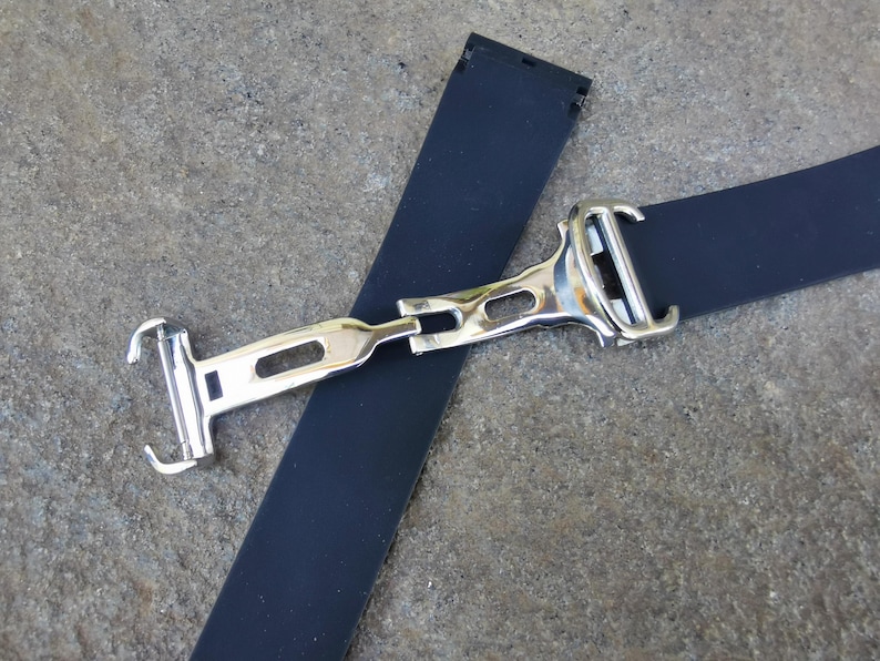 May include: A close-up of a watch strap with a silver-colored clasp. The strap is dark blue and made of a flexible material. The clasp is rectangular with a polished finish, and the strap is lying on a textured surface.