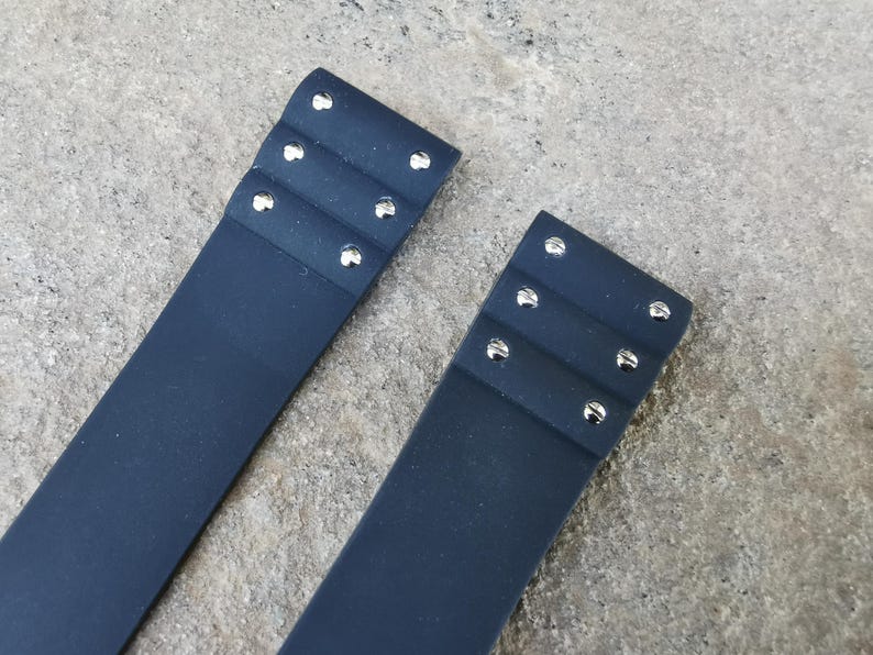 May include: Two navy blue leather straps with silver rivets. Each strap has three rows of rivets. The straps are on a textured, gray surface.