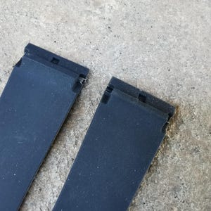 May include: Two black watch straps with a textured surface. Each strap has a rectangular shape with a connecting mechanism at the top. The straps are positioned against a gray, textured background.