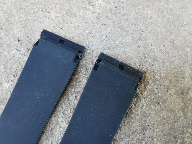 May include: Two black watch straps with a textured surface. Each strap has a rectangular shape with a connecting mechanism at the top. The straps are positioned against a gray, textured background.