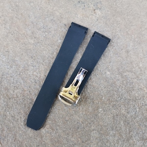 May include: Two-piece black watch strap with a silver and gold-toned clasp. The strap is made of a smooth, matte material. The clasp has a rectangular shape with a polished finish.
