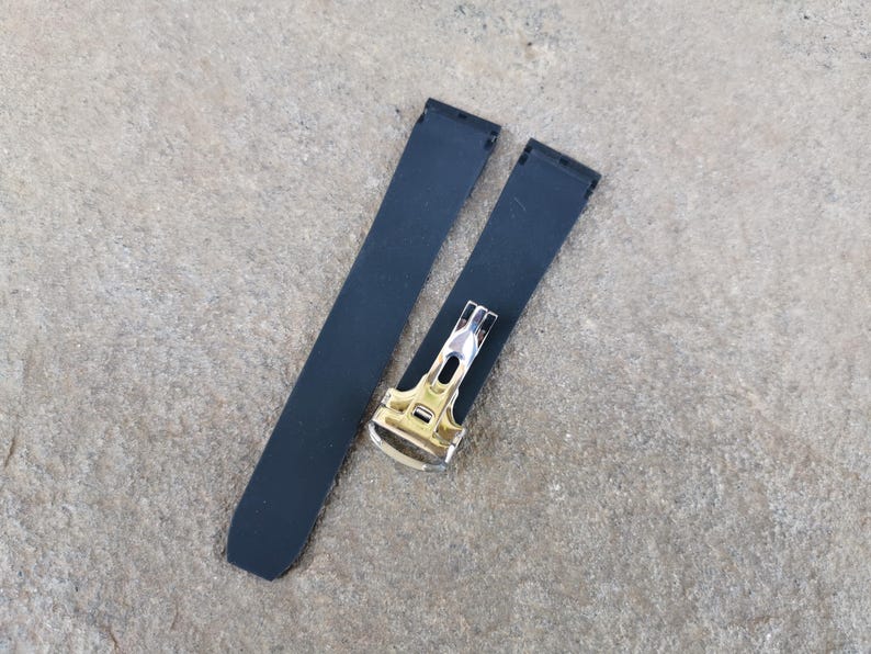 May include: Two-piece black watch strap with a silver and gold-toned clasp. The strap is made of a smooth, matte material. The clasp has a rectangular shape with a polished finish.