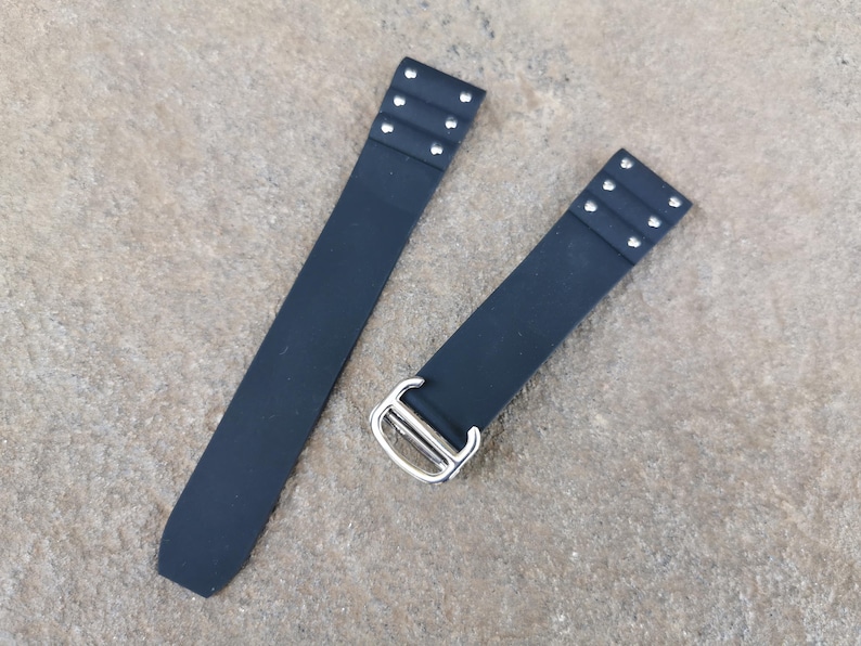 May include: Two-piece black watch strap with silver-tone hardware. Each strap section has three pairs of silver rivets. The strap has a rectangular shape and is made of a smooth, matte material.