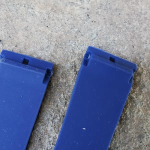 May include: Two vibrant blue watch straps are displayed against a textured, gray background. Each strap has a rectangular shape with a small, notched section at the top. The straps are made of a smooth, possibly rubbery material.