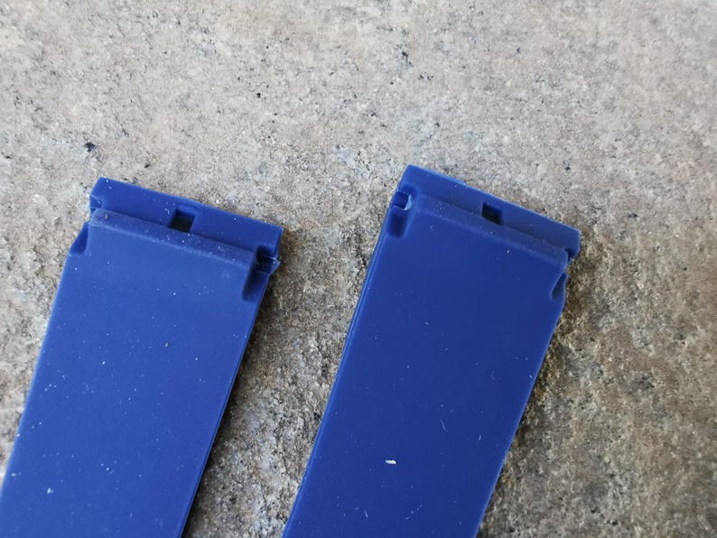 May include: Two vibrant blue watch straps are displayed against a textured, gray background. Each strap has a rectangular shape with a small, notched section at the top. The straps are made of a smooth, possibly rubbery material.