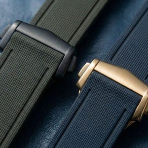 Könnte beinhalten: Nahaufnahme von zwei Uhrenarmbändern. Eines ist olivgrün mit einer schwarzen Schließe, das andere ist marineblau mit einer goldenen Schließe. Beide Armbänder haben eine strukturierte Oberfläche und sind vor blauem Hintergrund platziert.